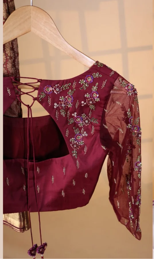 Maroon silk bridal blouse on a wooden hanger, hand-embroidered across the shoulder with multicoloured floral thread work, sequins and beading, with sheer net sleeves and a corded lace-up back finished in tassels