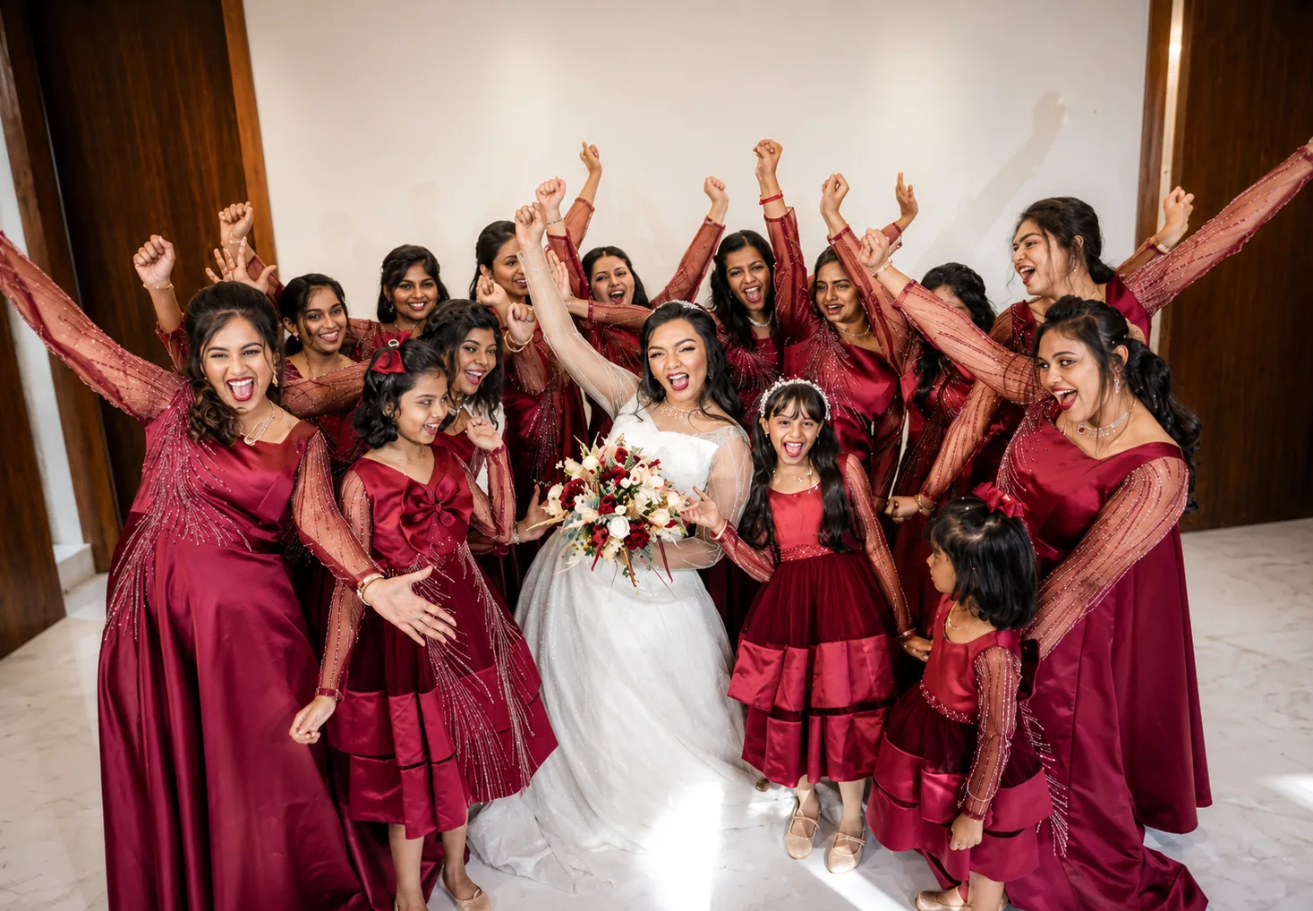 Bride in a white gown holding a red and ivory bouquet, cheering with her arms raised among a bridal party of women and young girls in matching wine-red satin gowns with sheer beadwork sleeves